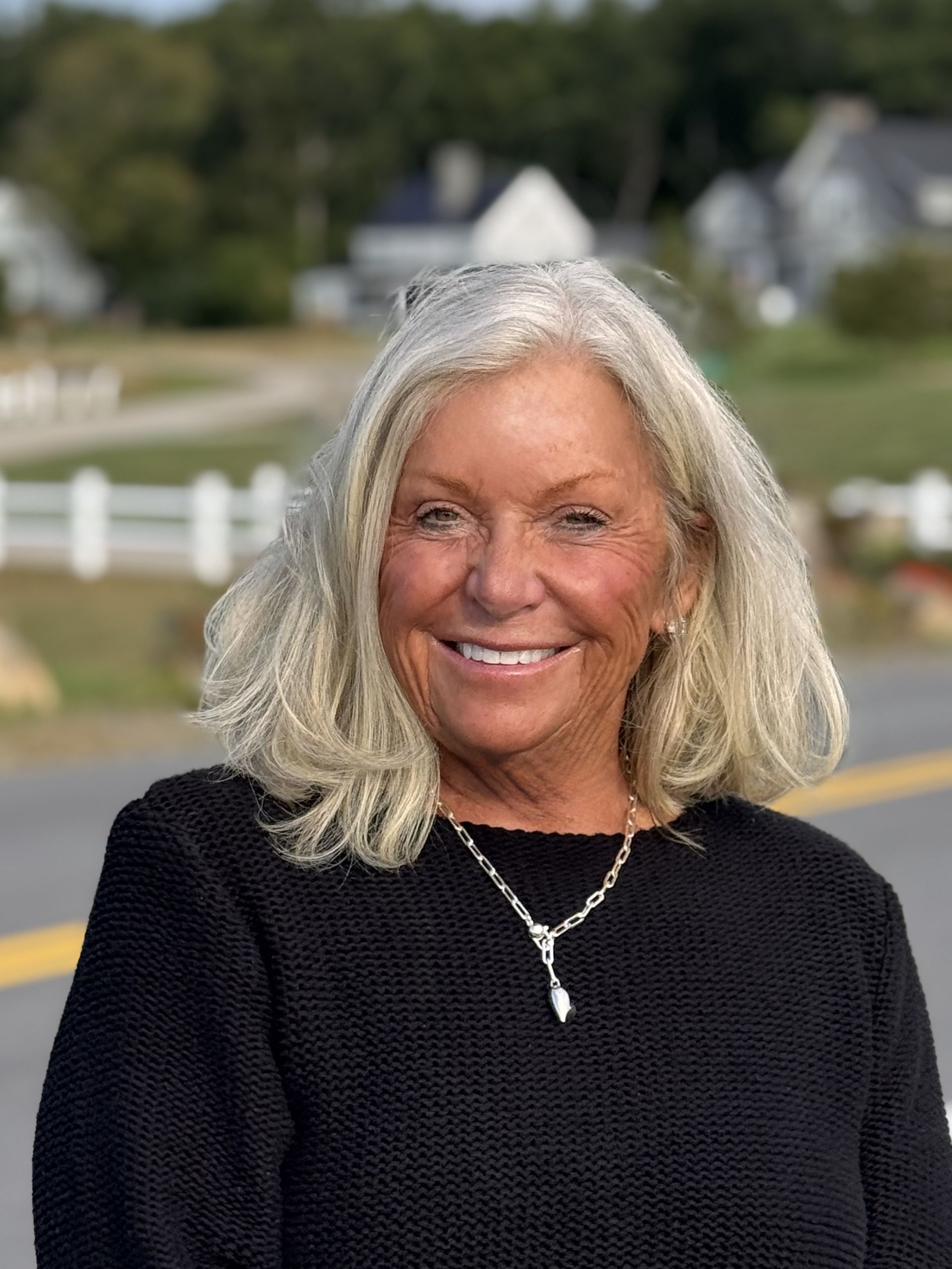 Smiling woman with gray hair in a black sweater, standing outdoors near a white fence and houses
