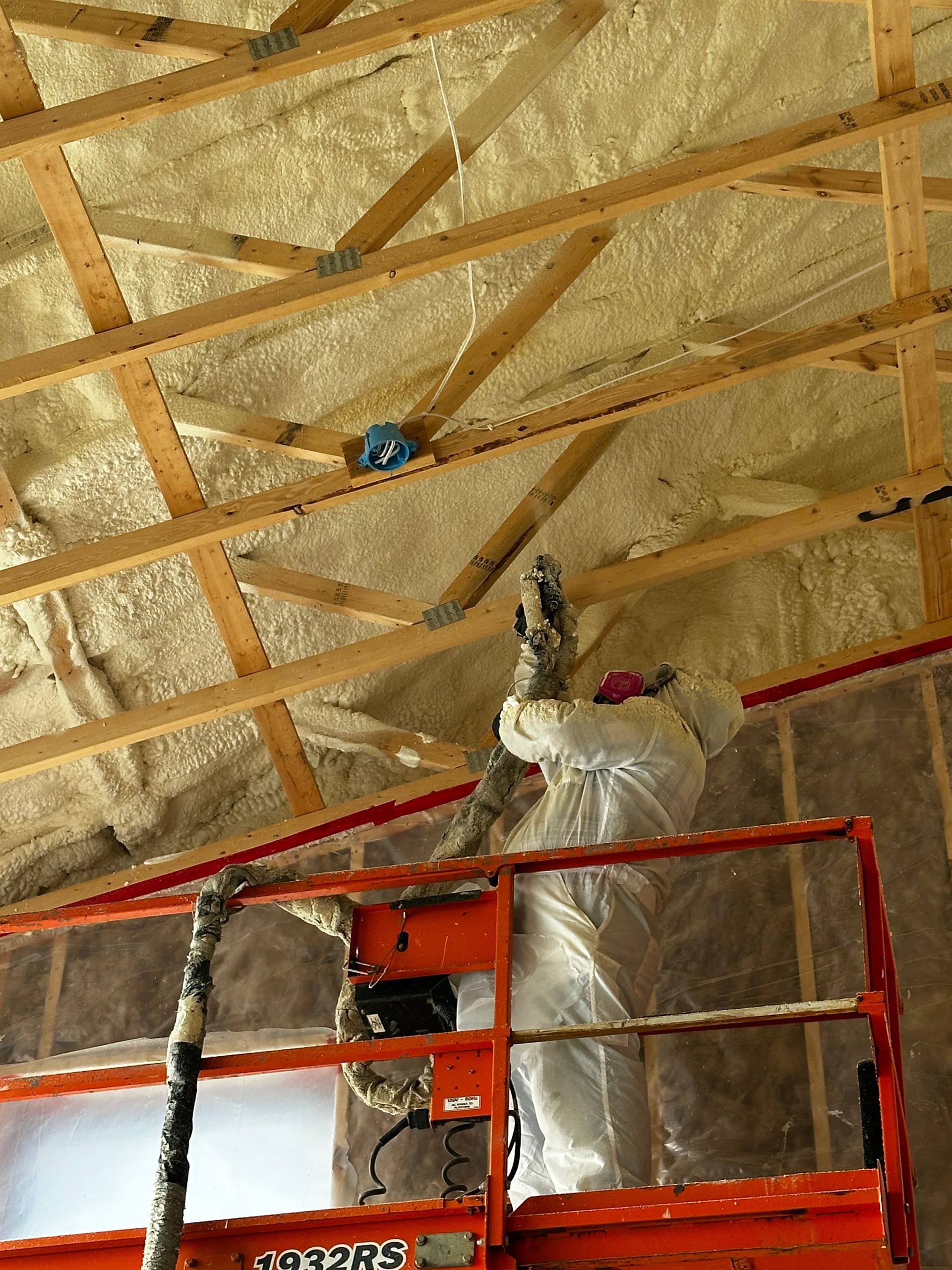 Sprayed insulation filling wall studs; light tan foam being applied. A worker in protective gear on a lift sprays foam insulation onto the wooden rafters of a building ceiling.