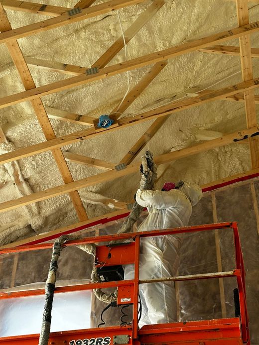 Sprayed insulation filling wall studs; light tan foam being applied. A worker in protective gear on a lift sprays foam insulation onto the wooden rafters of a building ceiling.