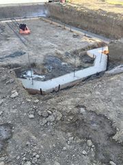 Construction excavation with concrete foundation walls and a small red machine in a dirt pit.