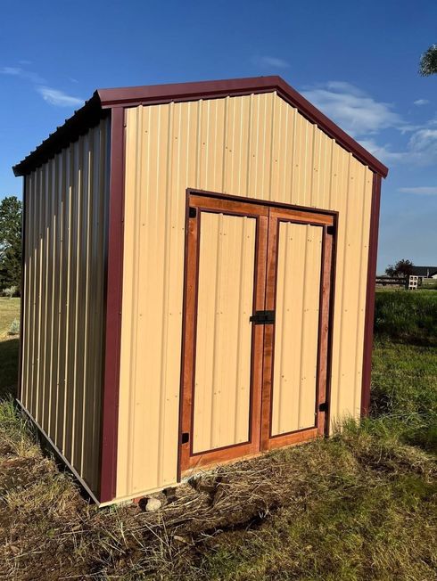 Small wooden shed with tan siding, red trim, and double doors standing in a grassy field under a blue sky