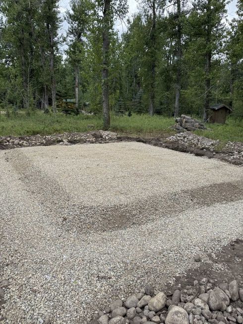 Freshly leveled gravel pad in a wooded area, bordered by rocks and trees.