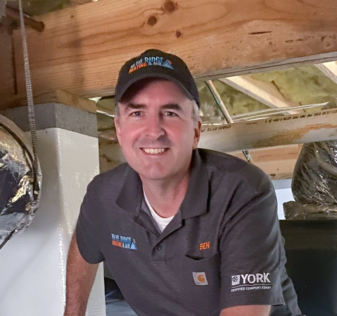 Man in a cap and polo shirt smiles while inspecting ductwork. Setting appears to be a crawl space.
