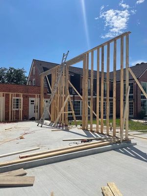Wood-framed house under construction in a driveway, with tall wall framing and ladders in bright sunlight