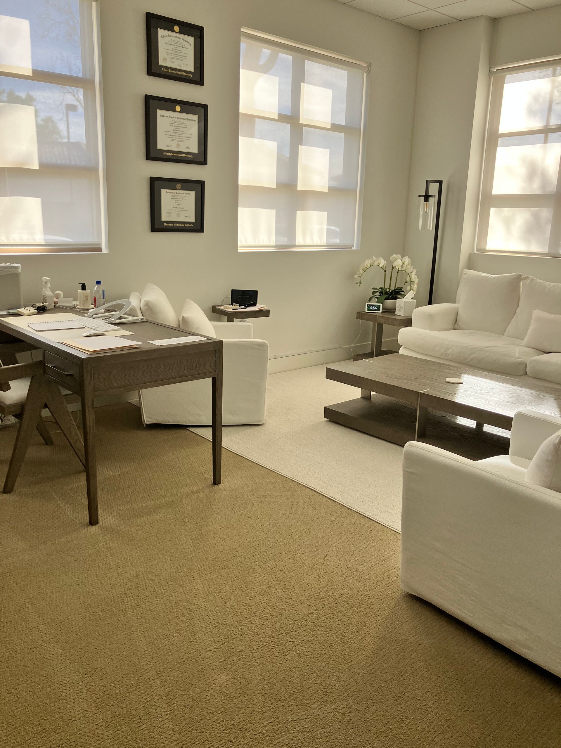 Bright living room with white furniture, a desk, framed wall art, and large sunlit windows.