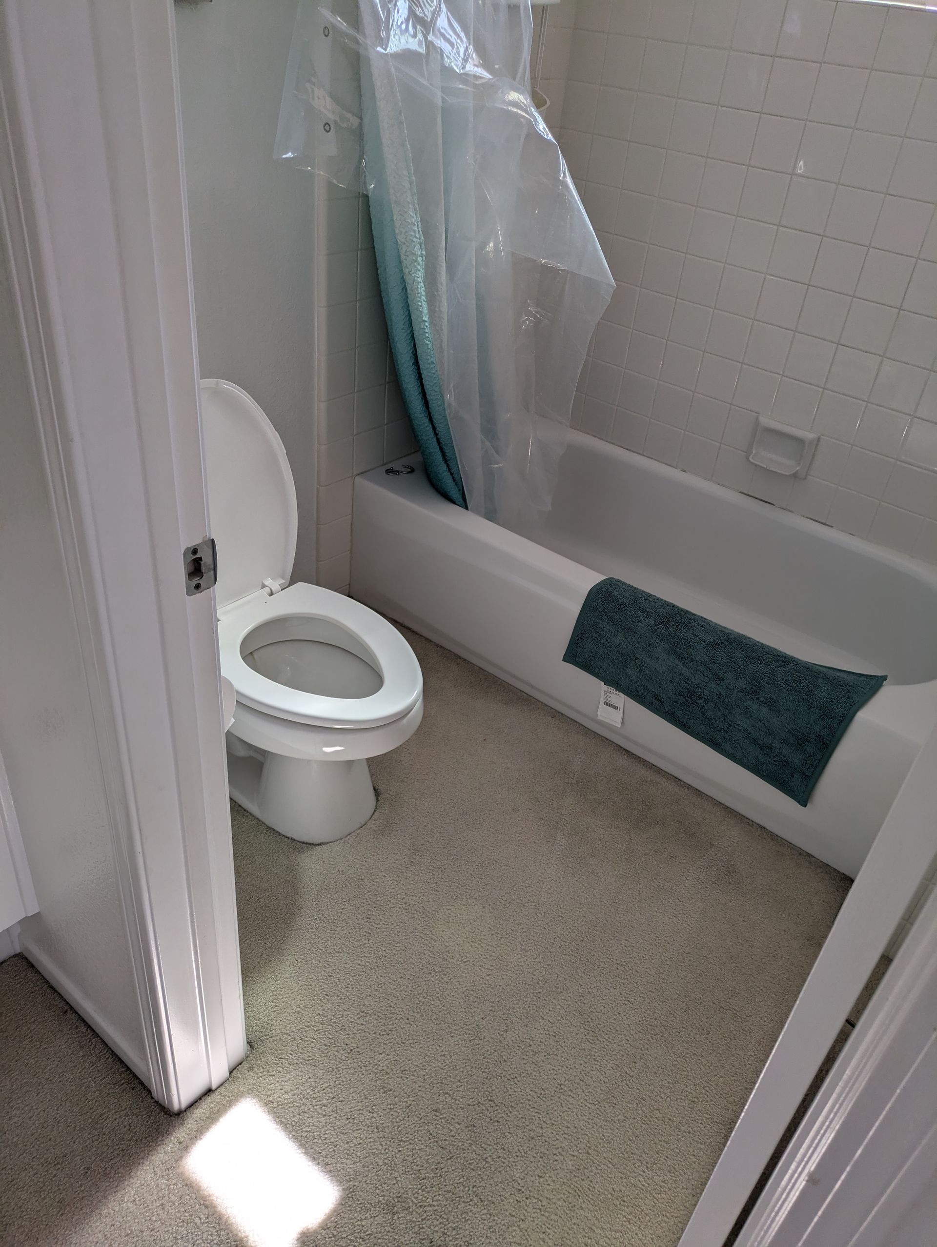 Small bathroom with open toilet, bathtub, and shower curtain; teal towels hang on the tub edge.