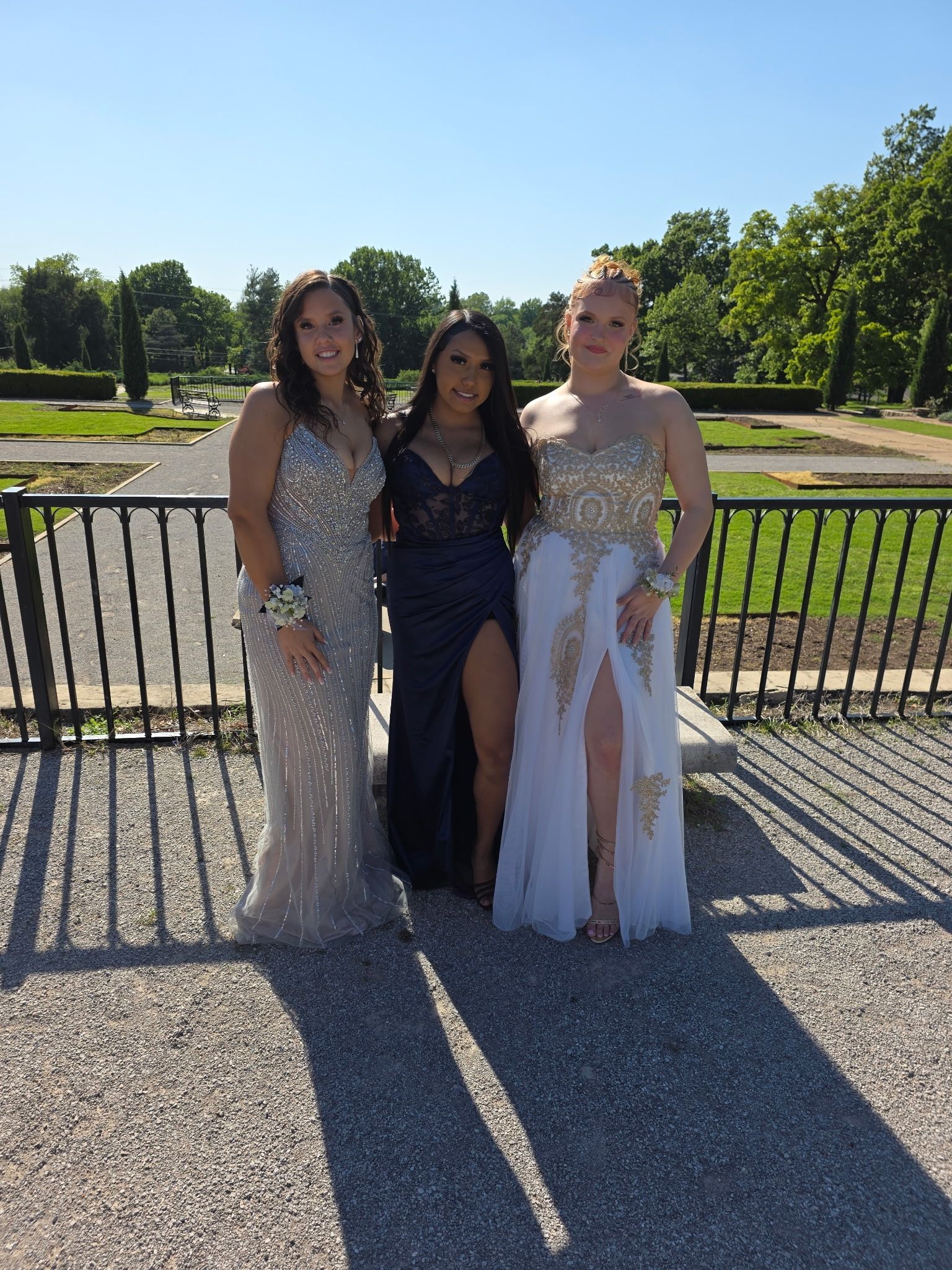 Three people posing outdoors in formal dresses by a black fence on a sunny day.