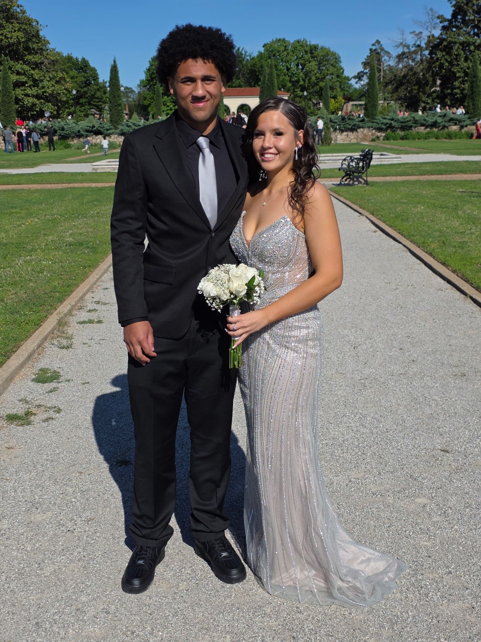 Couple posing on a park path, smiling; one holds a bouquet, both dressed in formal attire.