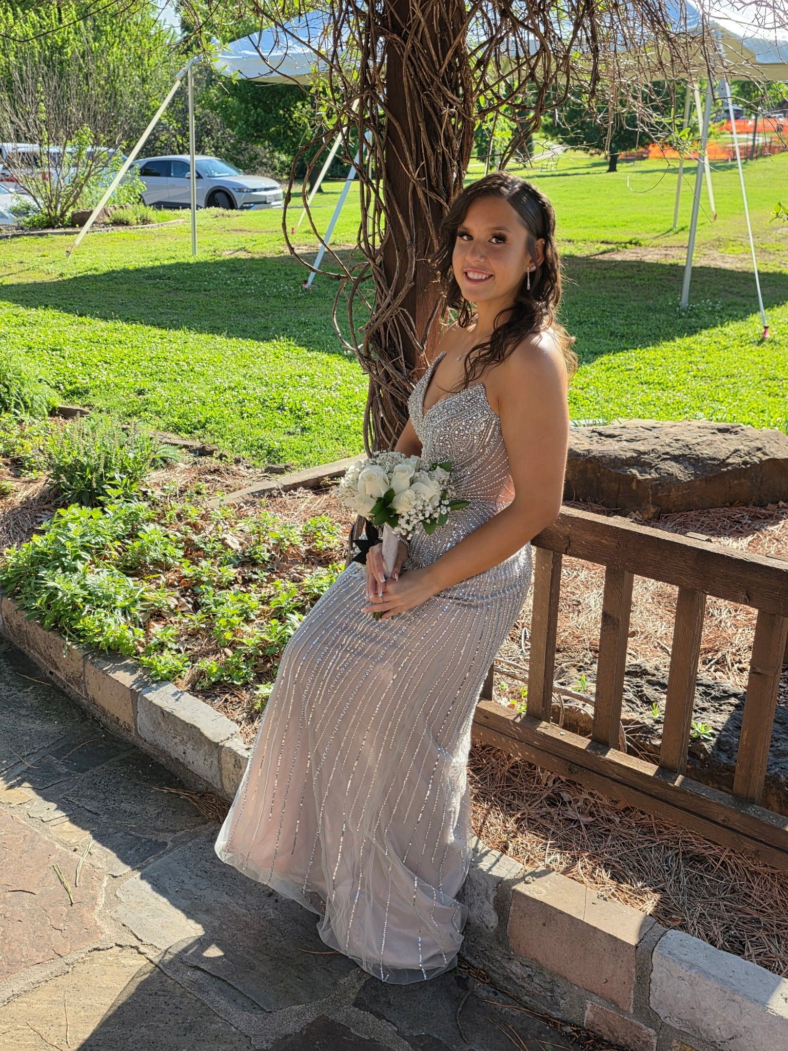 Woman in a sparkling dress sitting on a wooden bench in a sunny garden with green grass and flowers