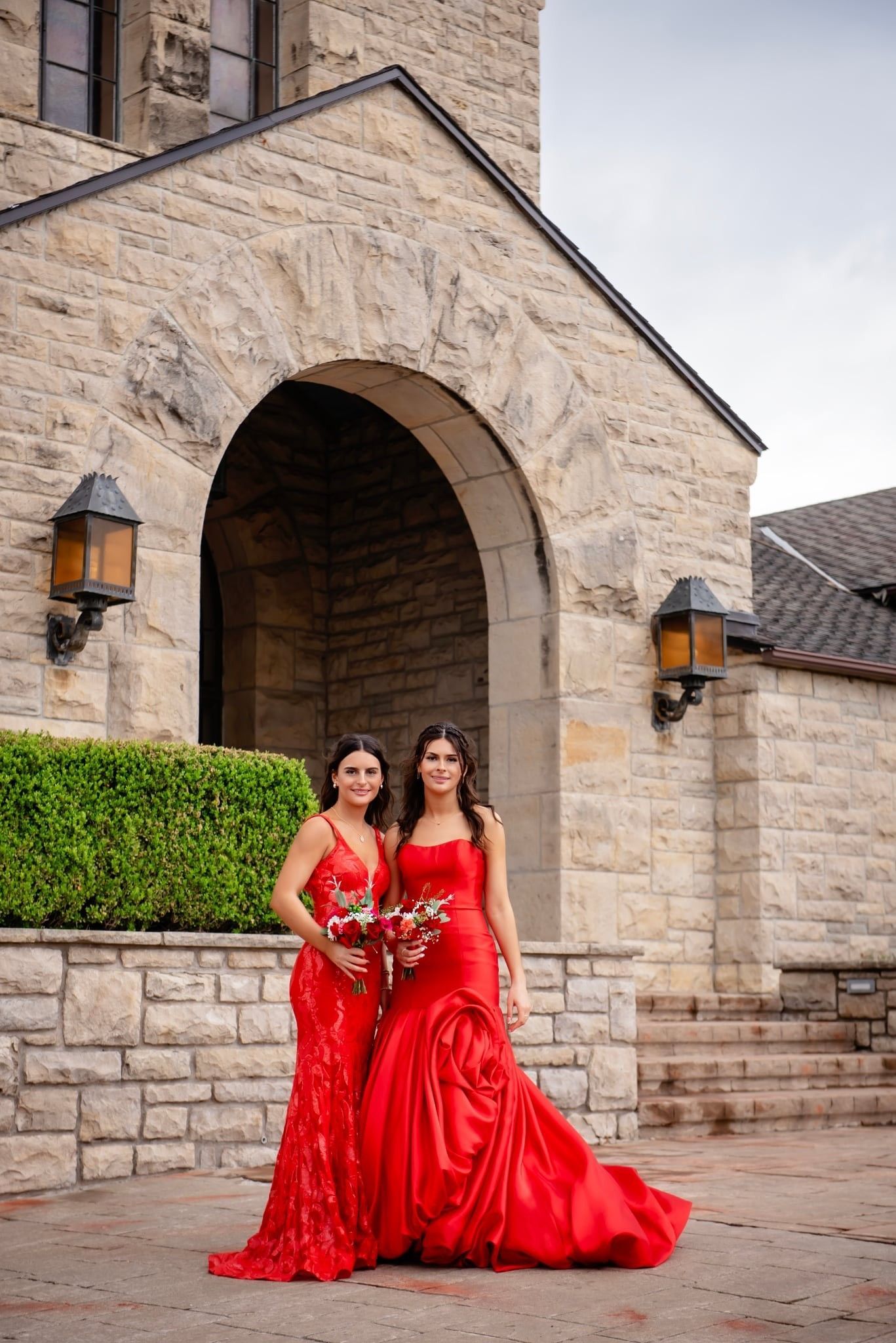 Two women in matching red gowns posing by a stone archway and wall