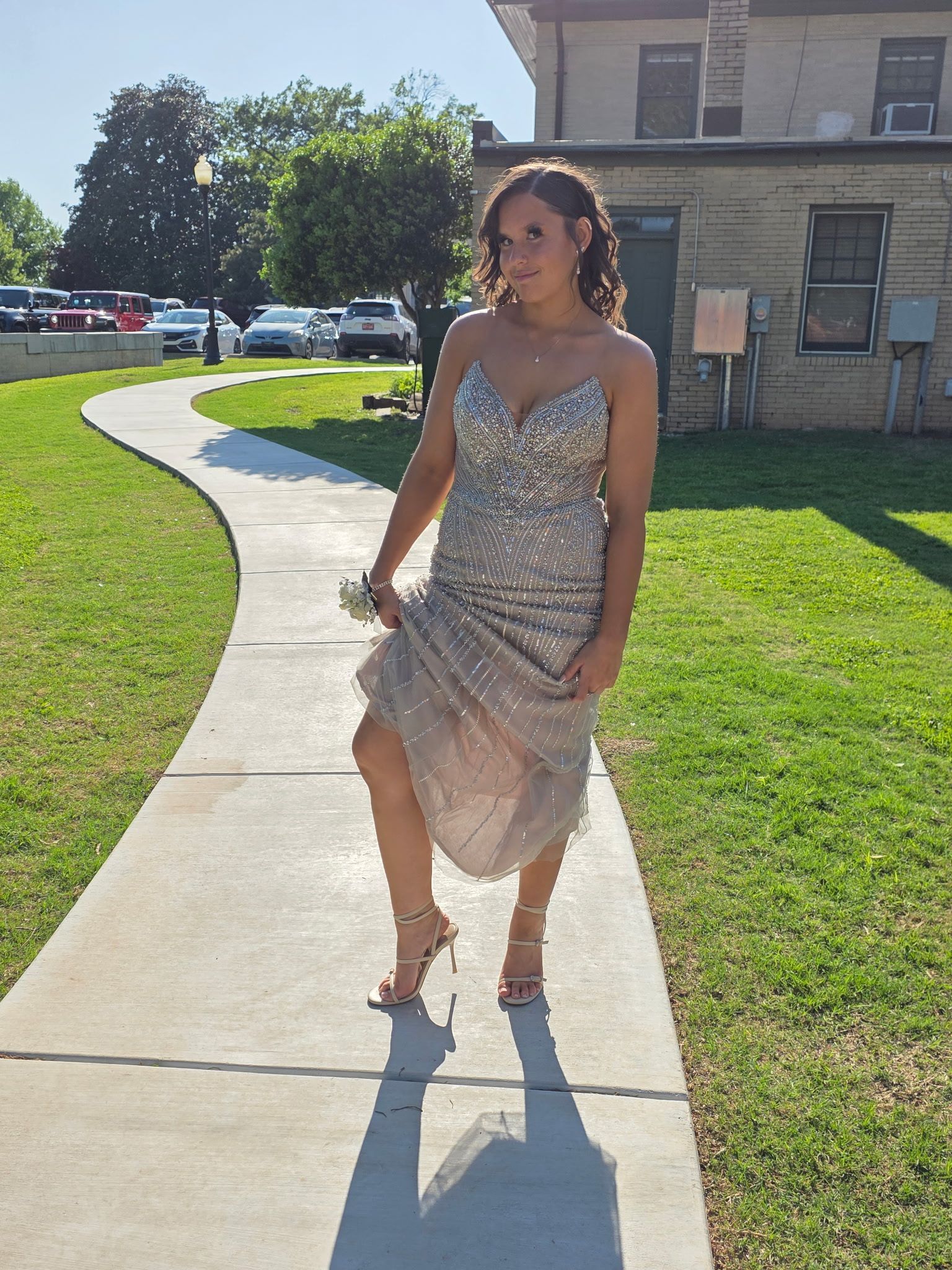 Person in a sparkly dress walking on a sunlit sidewalk beside a grassy lawn and building.