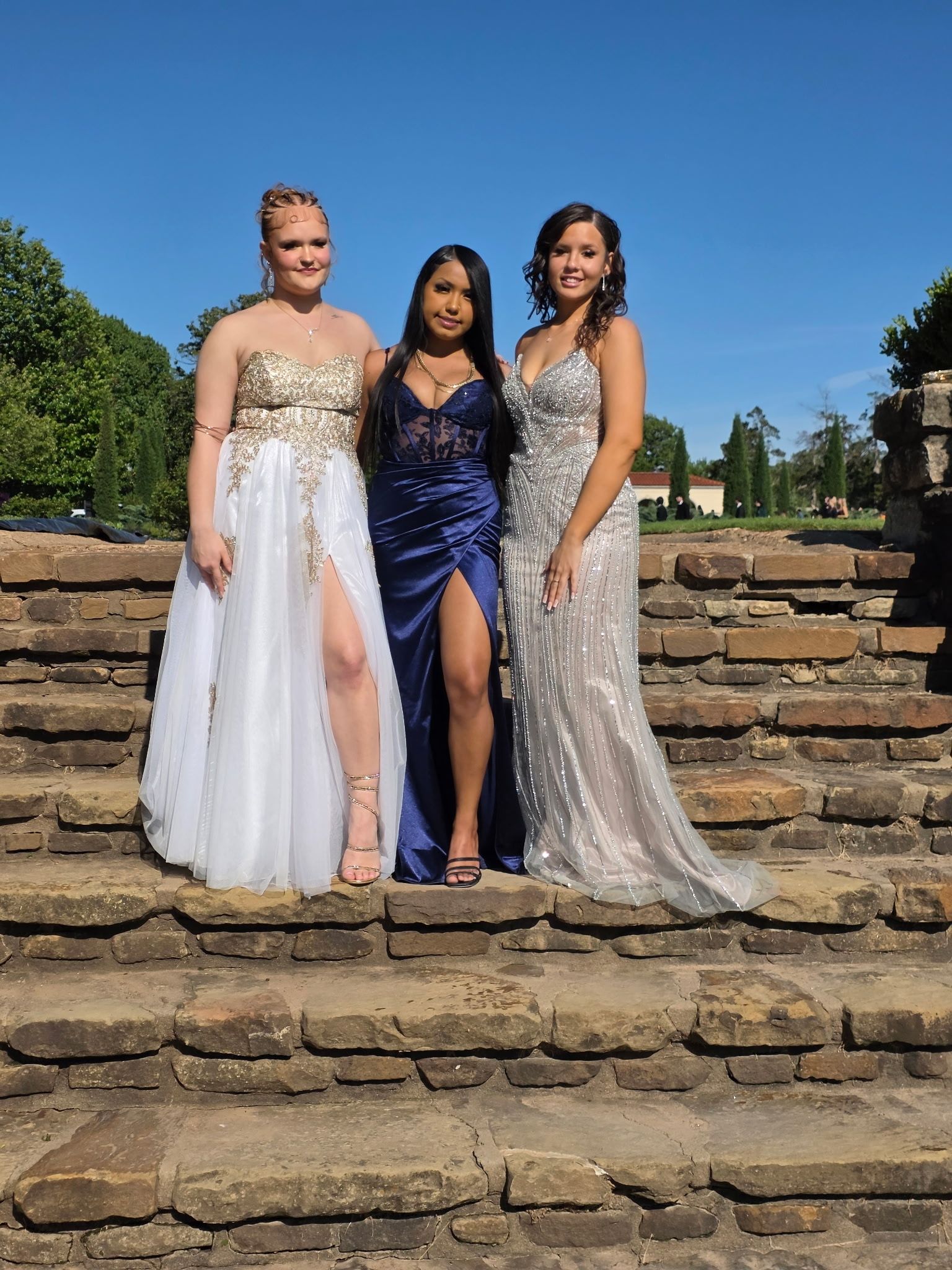 Three women in formal dresses posing on stone steps under a clear blue sky.