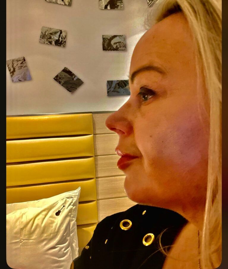 Side-profile of a blonde woman seated indoors beside a yellow upholstered headboard and wall photos.