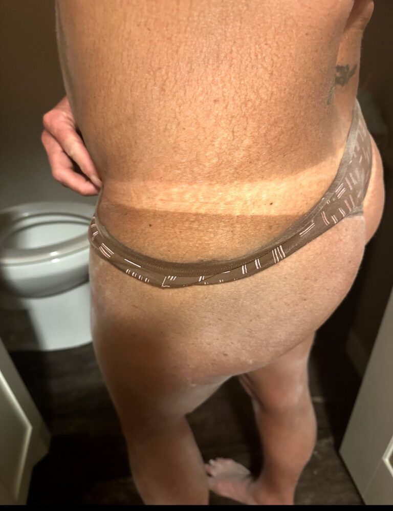 Rear view of a person standing in a bathroom, wearing a brown thong and showing a tan line