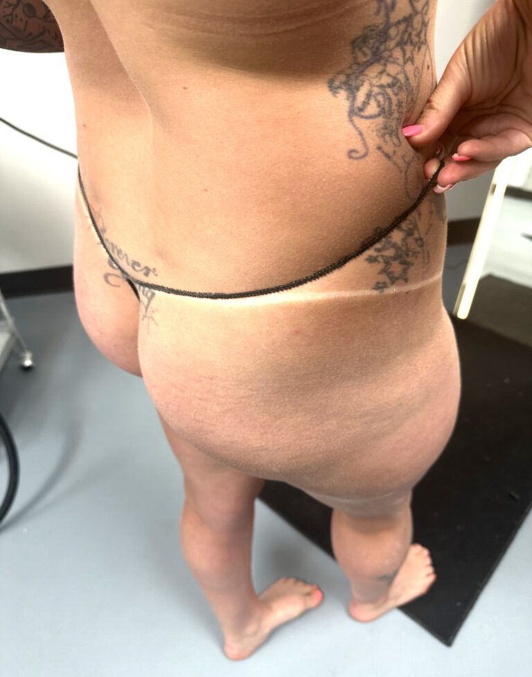 Rear view of a person in beige underwear with tattoos, standing indoors on a black floor mat.