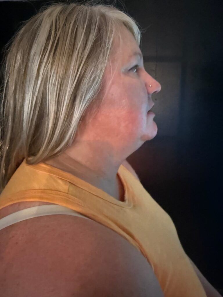 Side profile of a woman in a sleeveless orange top, looking ahead in a dimly lit indoor setting.
