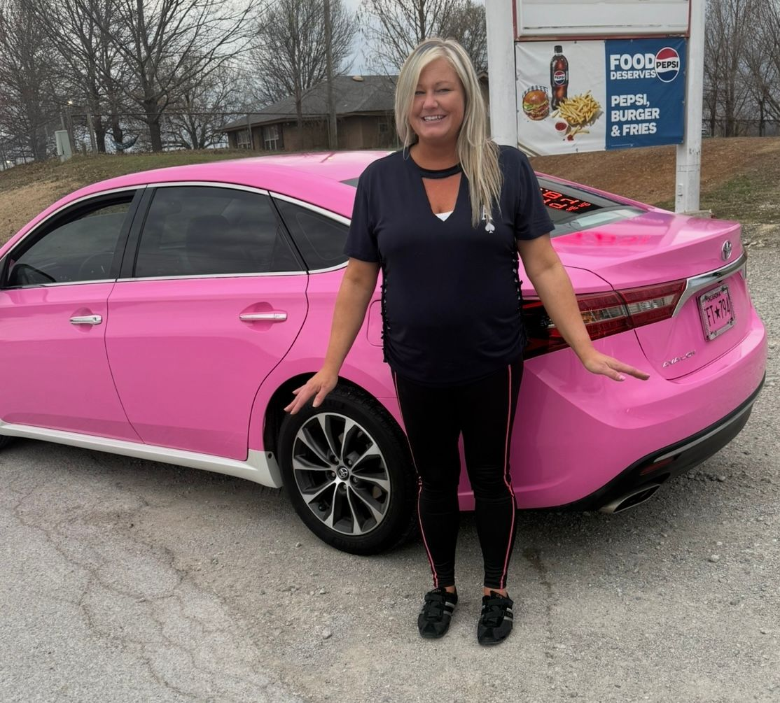 Woman standing beside a bright pink sedan in a parking lot, smiling at the camera.