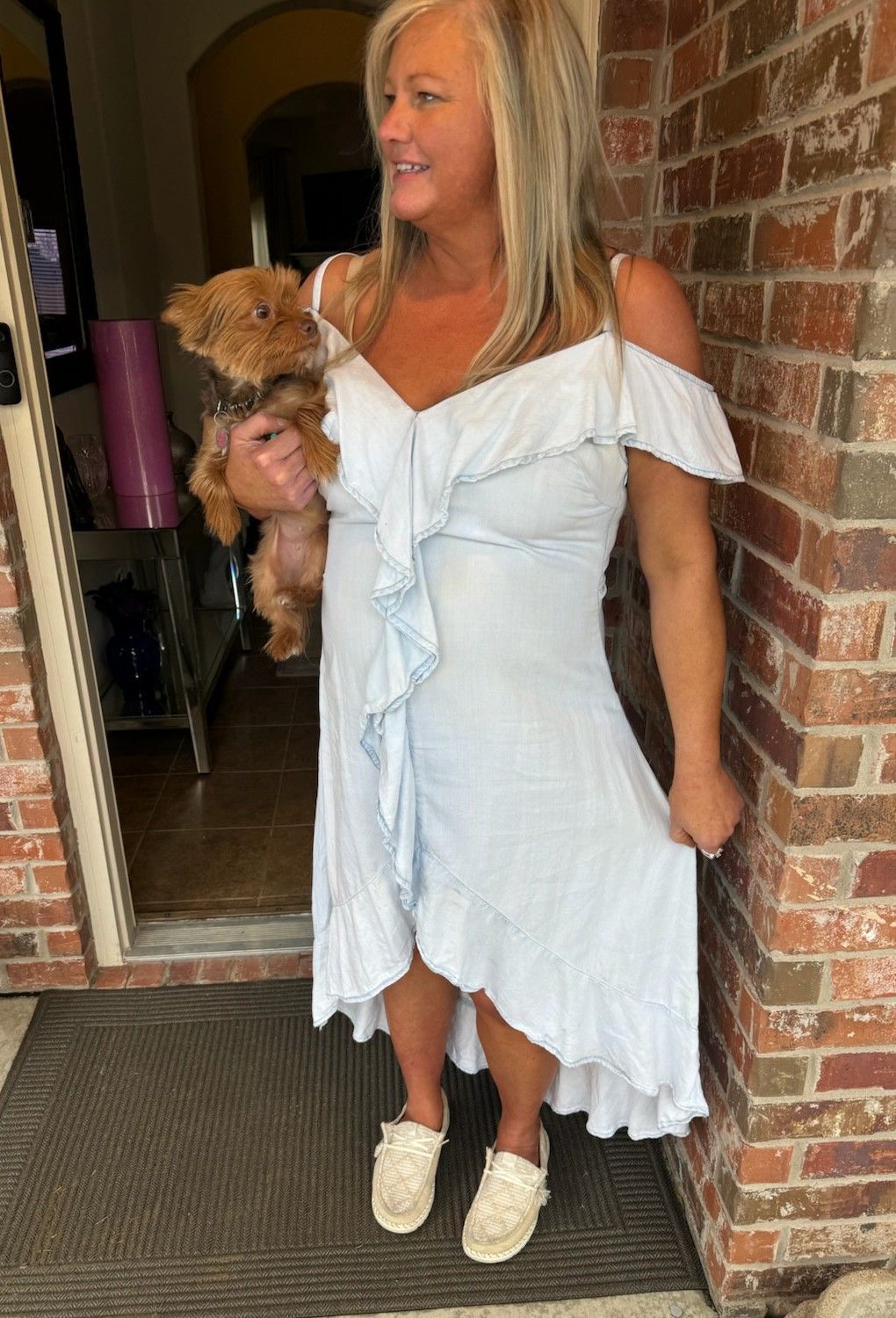 Woman in a white dress holding a small dog, standing by a brick doorway.