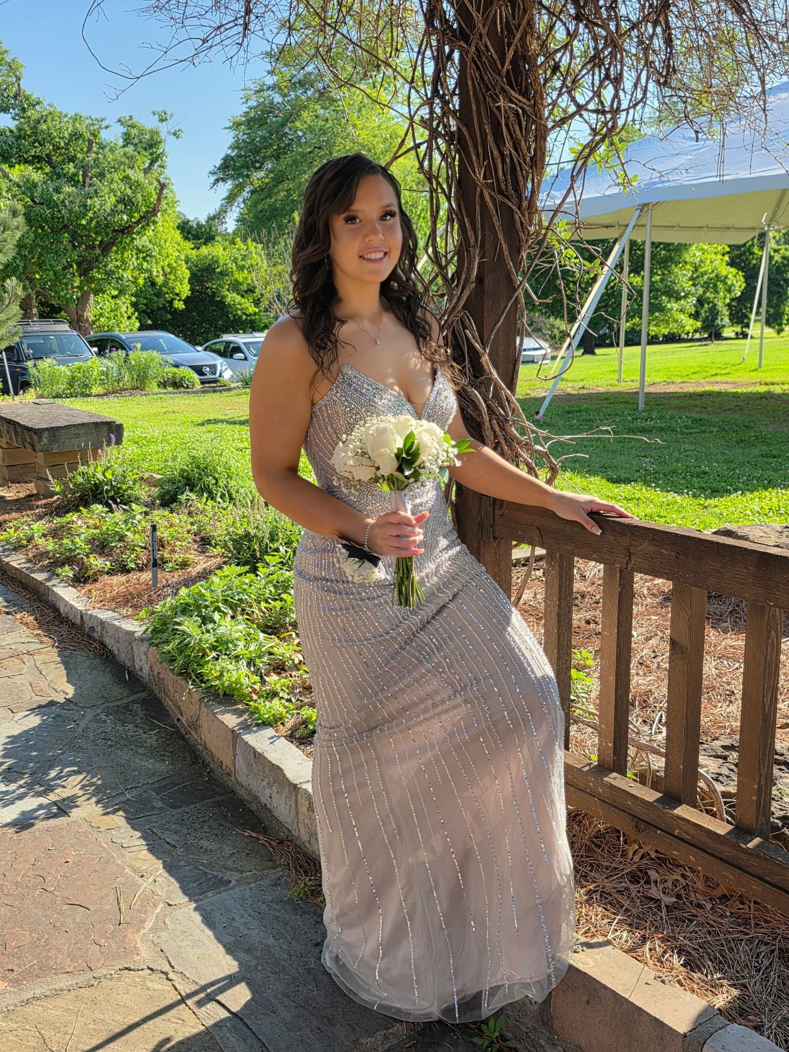 Person in a light pink dress holding a bouquet, standing by a wooden railing in a sunny garden.