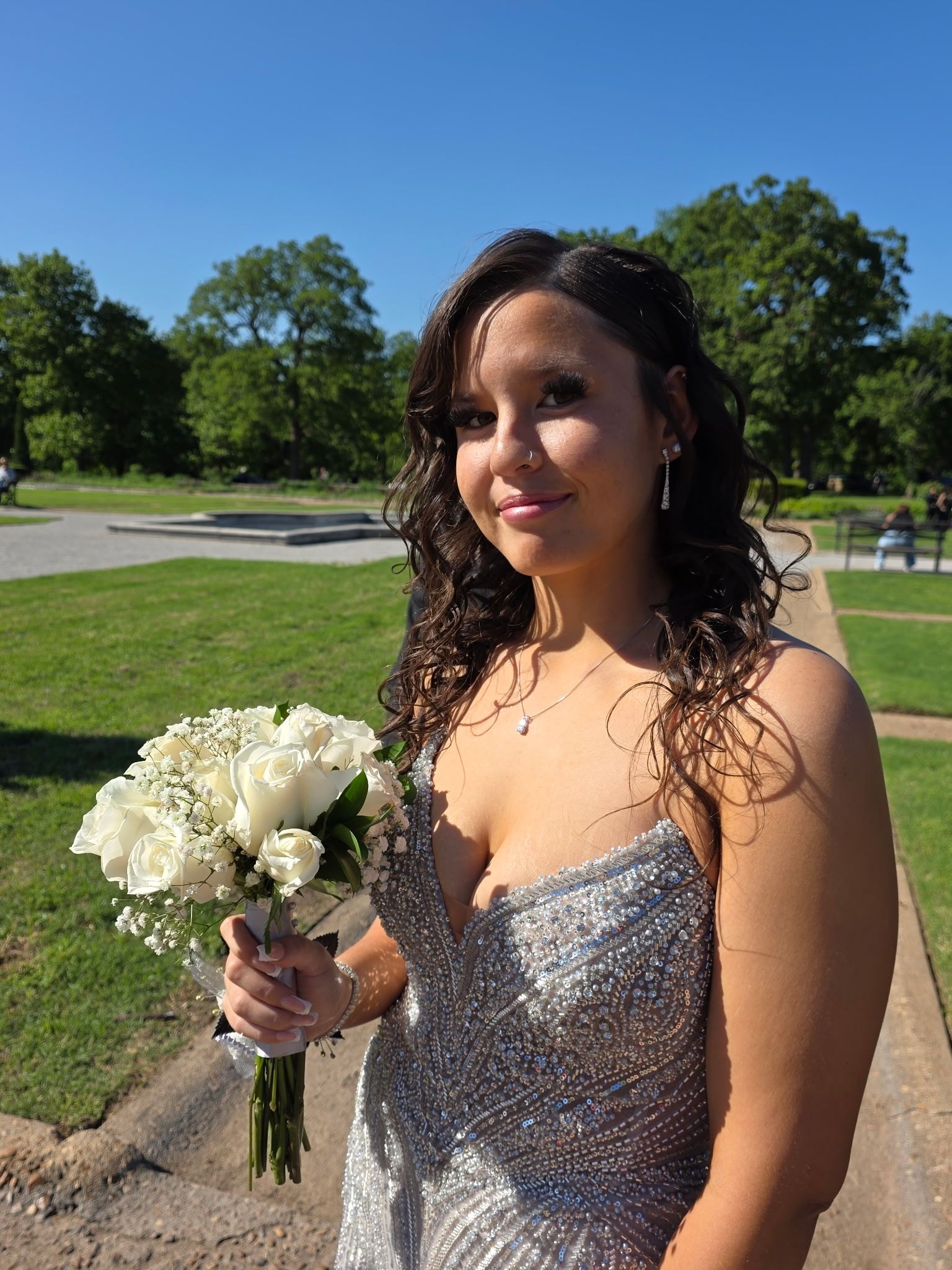 Woman in a silver dress holding a white bouquet outdoors on a sunny lawn.