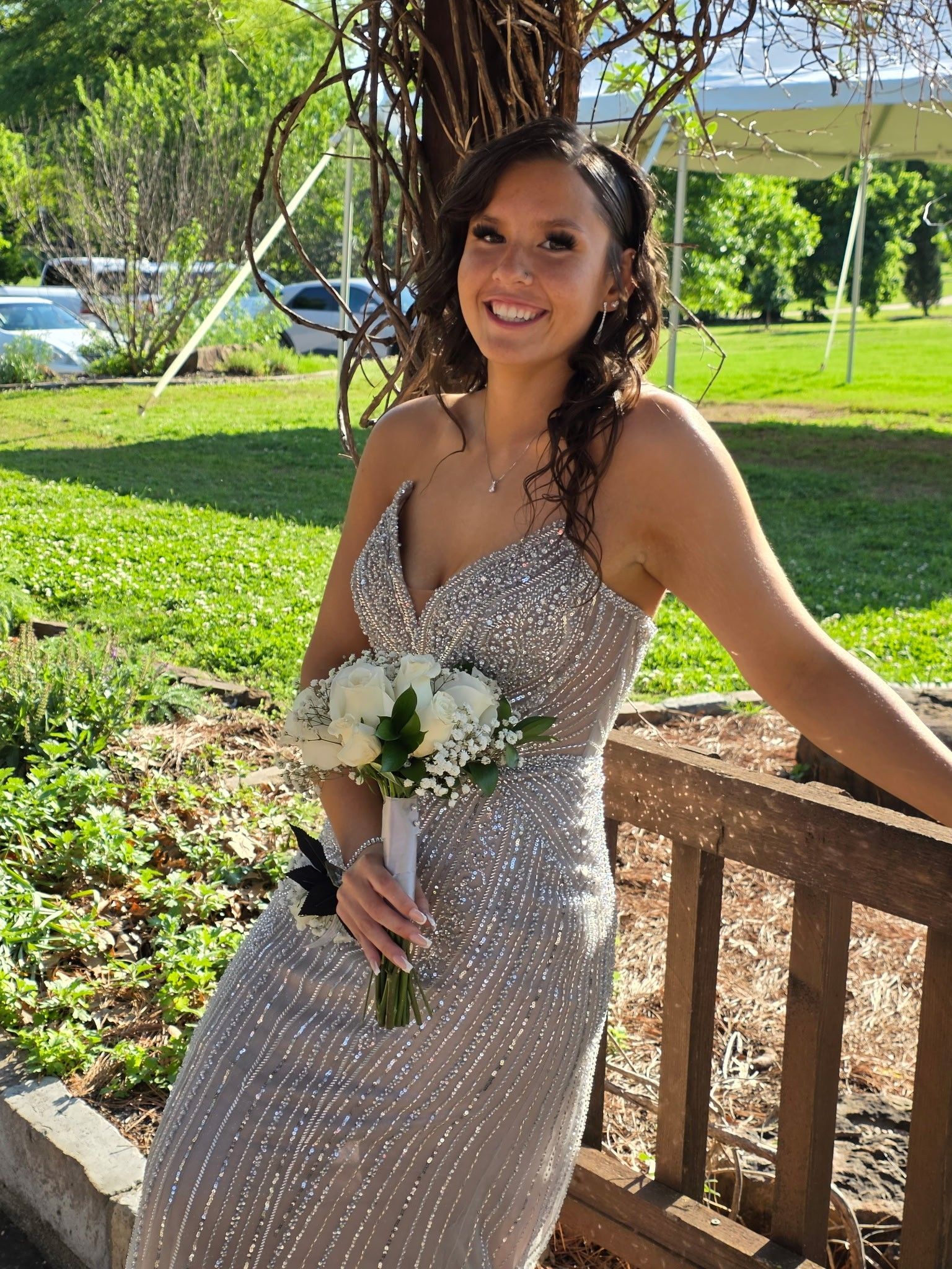 Woman in a sparkling silver dress holding a bouquet, smiling outdoors on a wooden porch.