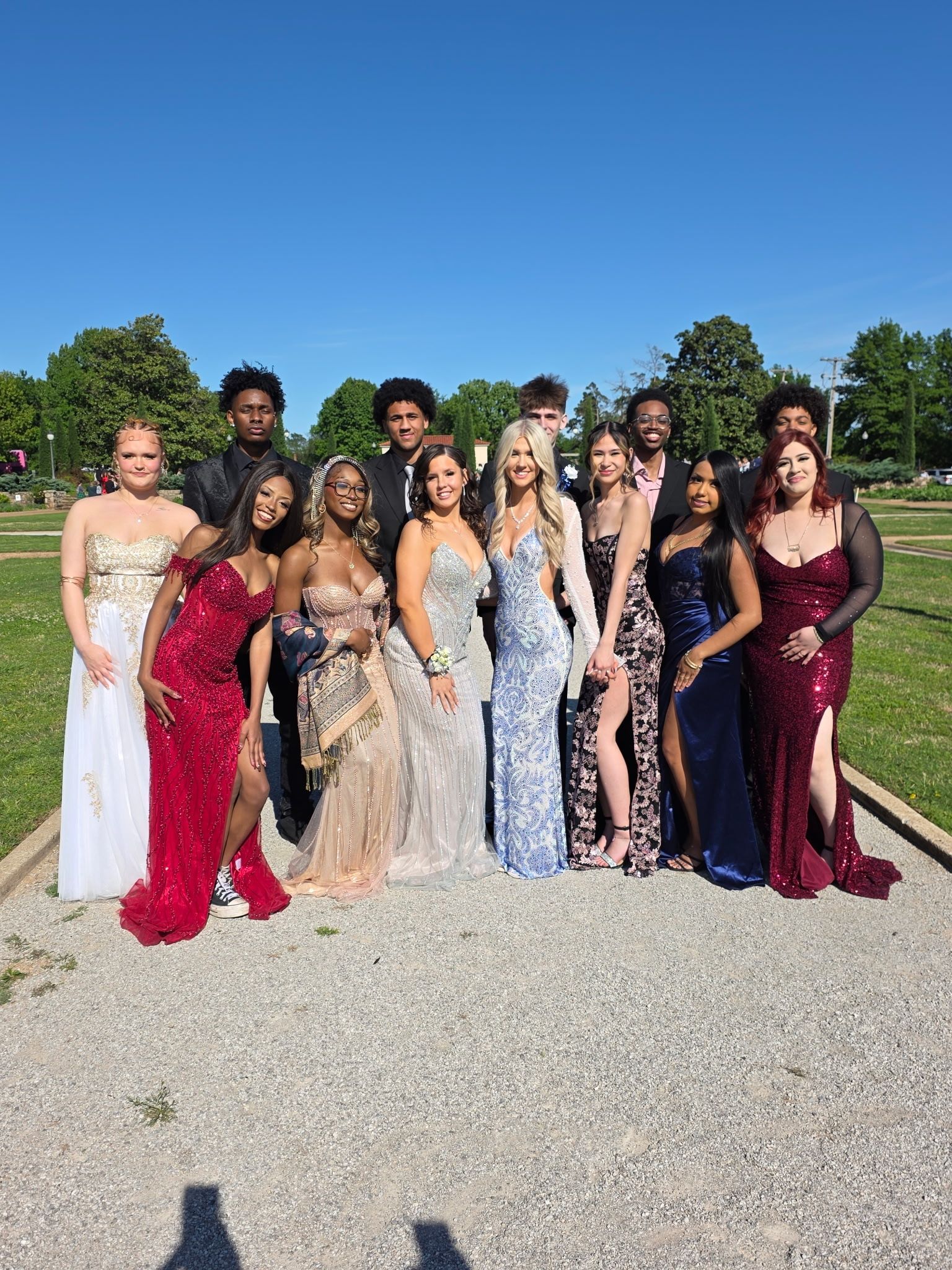 Group of people in formal dresses and suits posing outdoors on a gravel path under a clear blue sky