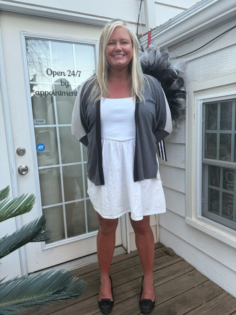 Woman standing on a porch in a white dress and gray cardigan, smiling beside a glass door.