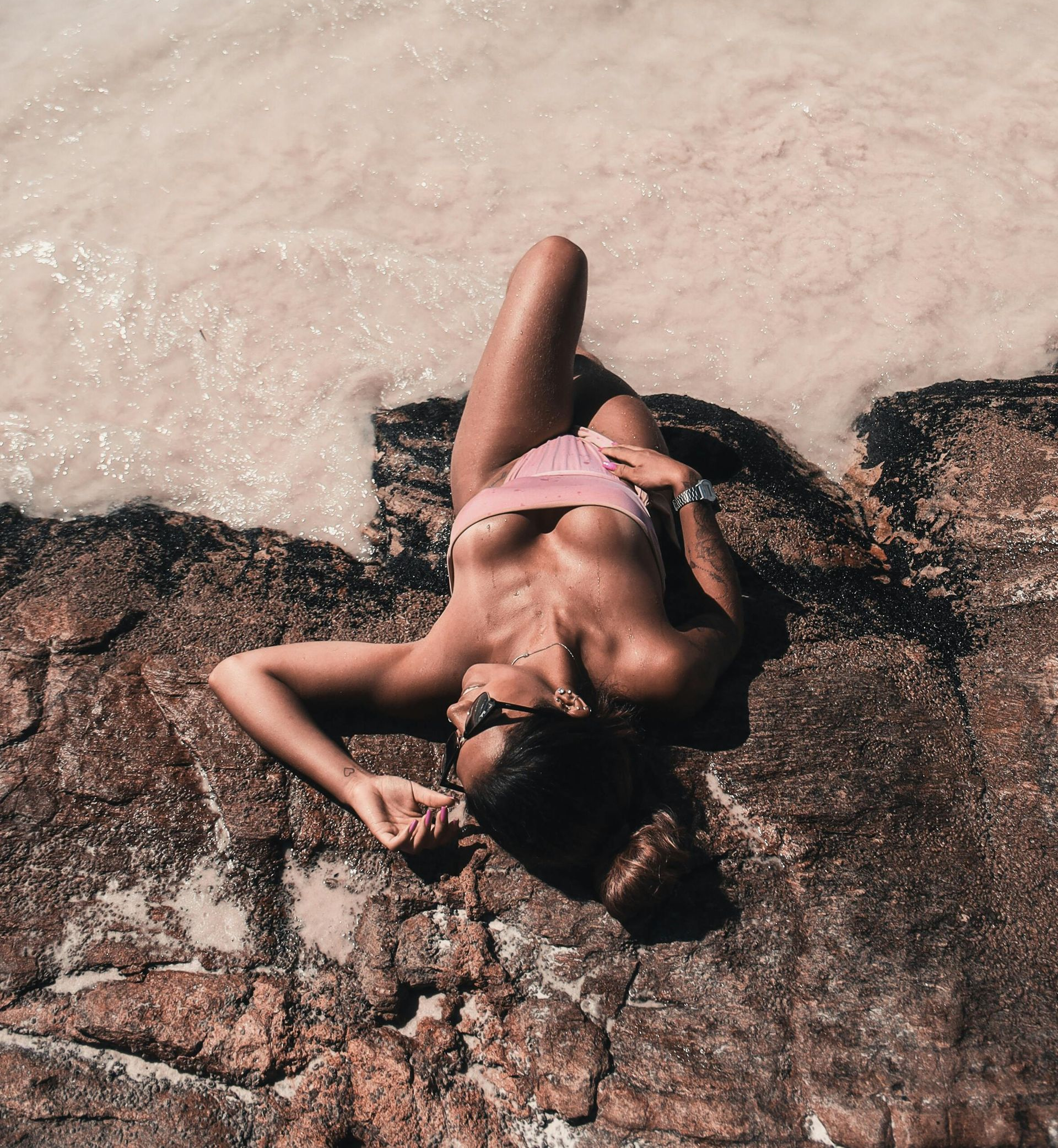 Person in a pink bikini lying on sunlit rocks beside foamy surf.