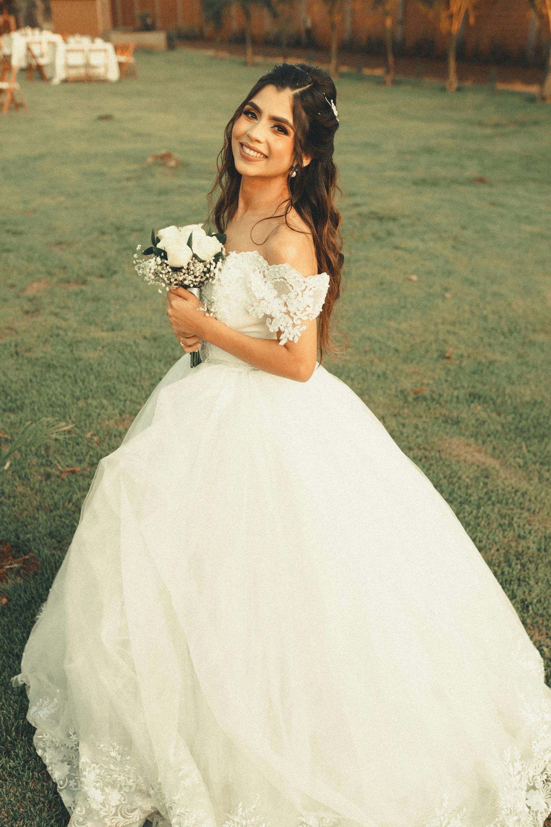 Woman in a white wedding dress holding a bouquet, smiling on a grassy lawn.