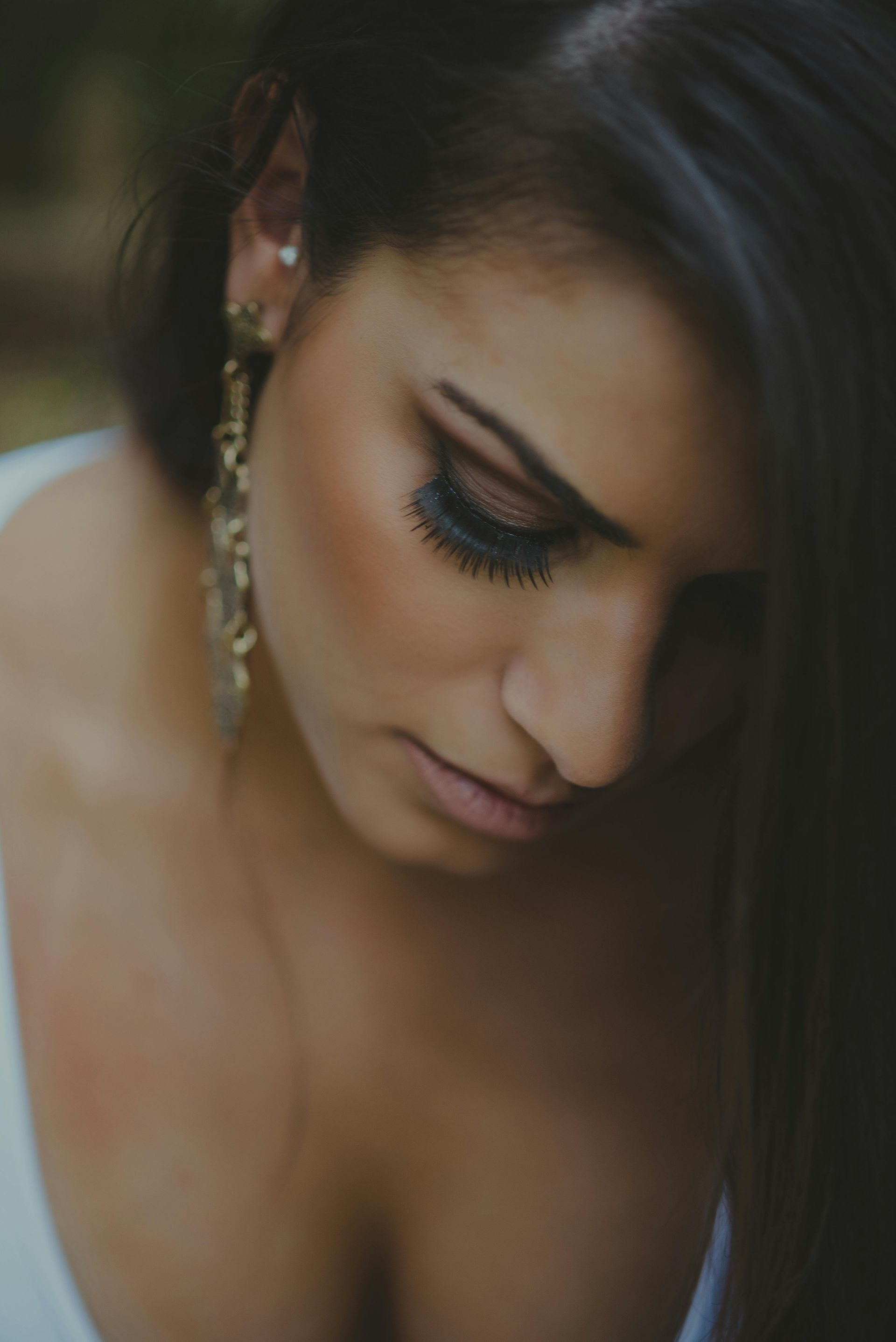 Close-up of a woman looking down, wearing long earrings and a white dress in soft light