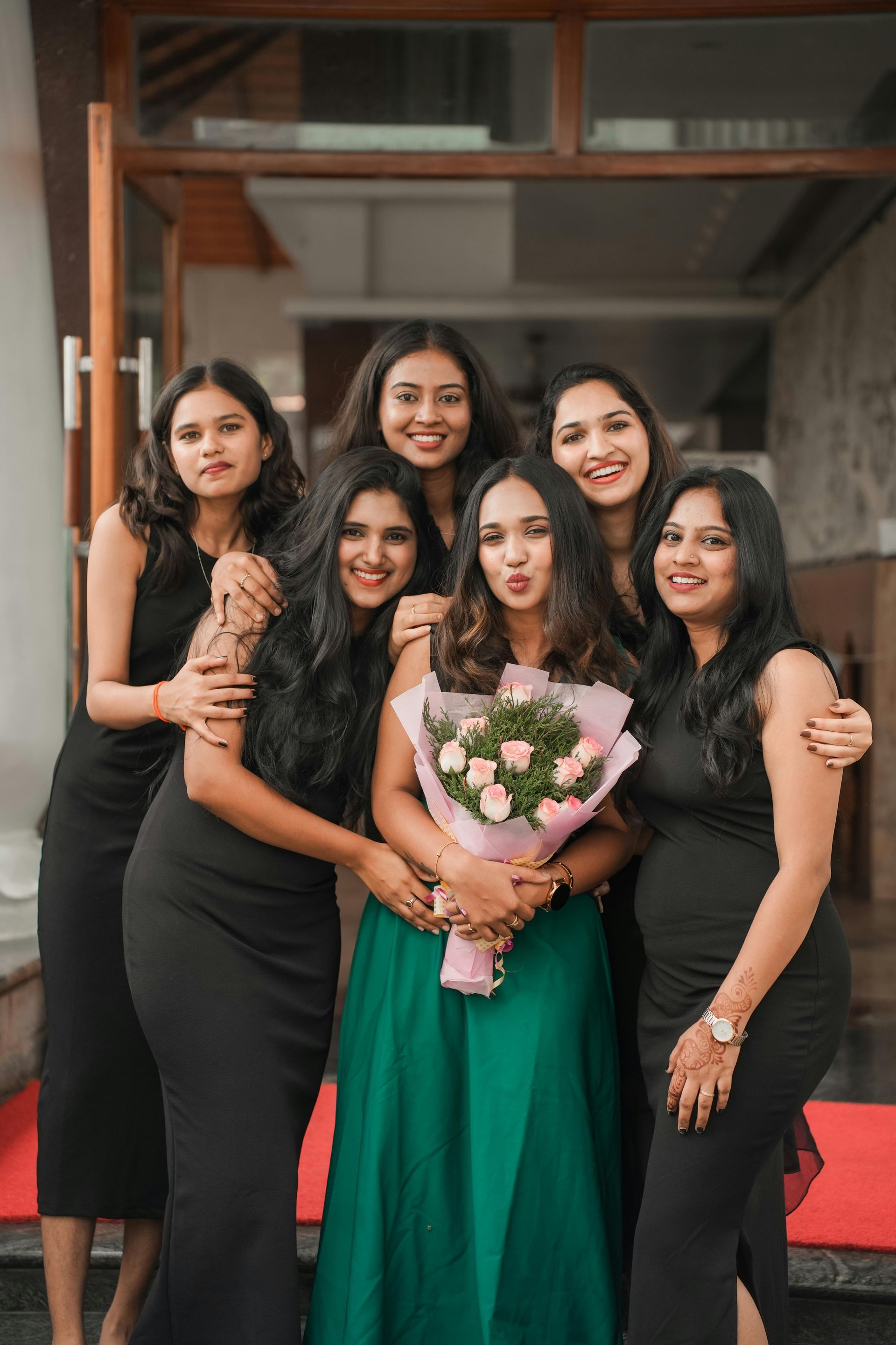 Five people in black and green dresses posing indoors, one holding a bouquet of flowers.