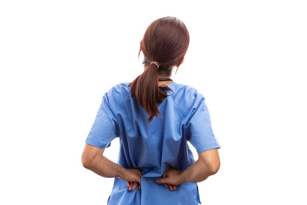 Nurse in blue scrubs, facing away, holding lower back, likely experiencing pain.