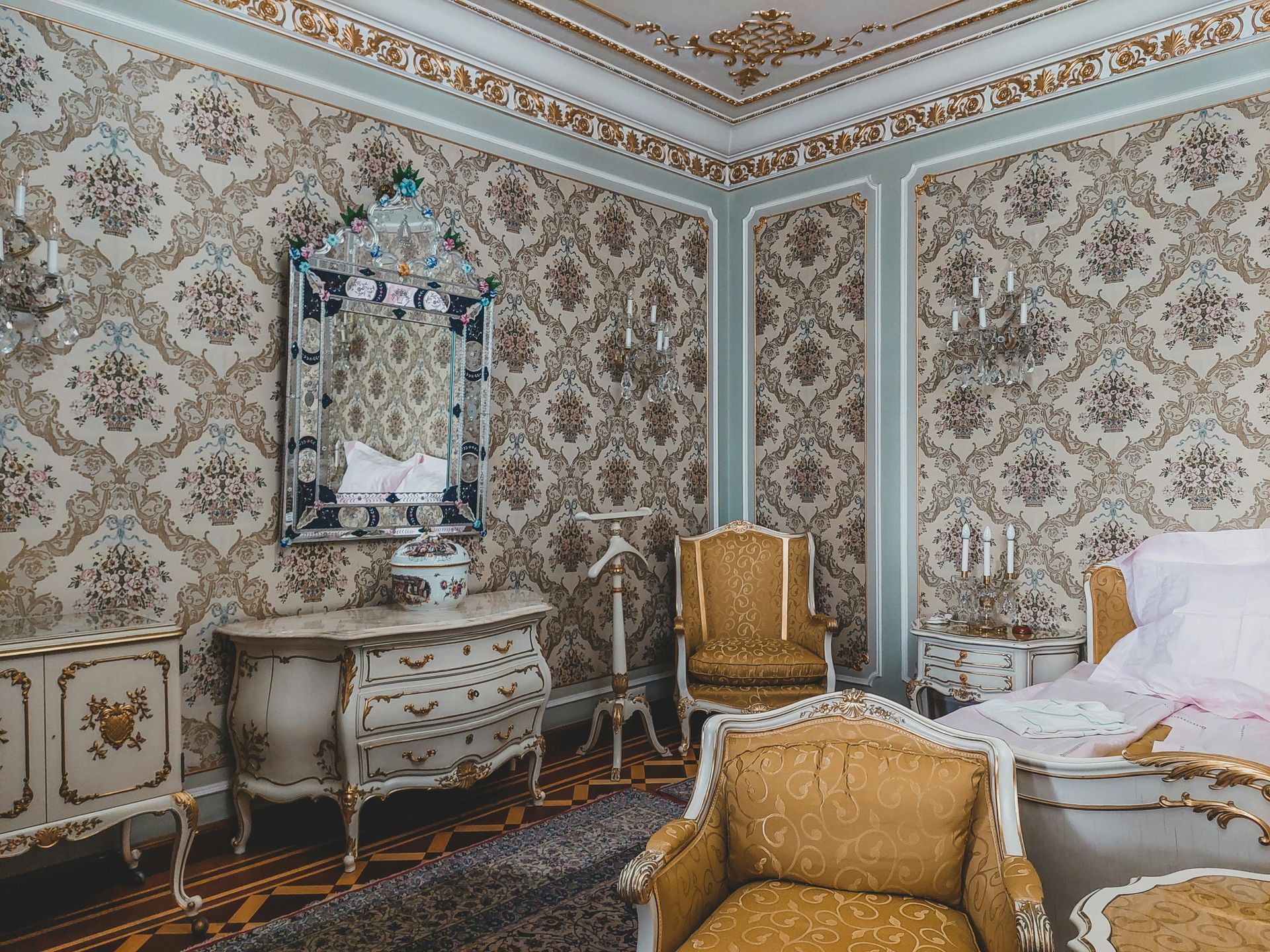 Ornate bedroom with patterned wallpaper, gilded trim, antique furniture, and a large mirror.