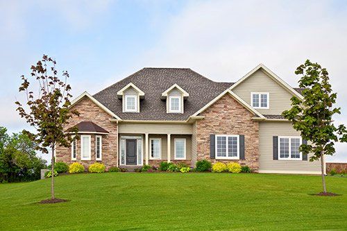 Suburban house with gray shingles, beige siding, front porch, and green lawn with trees
