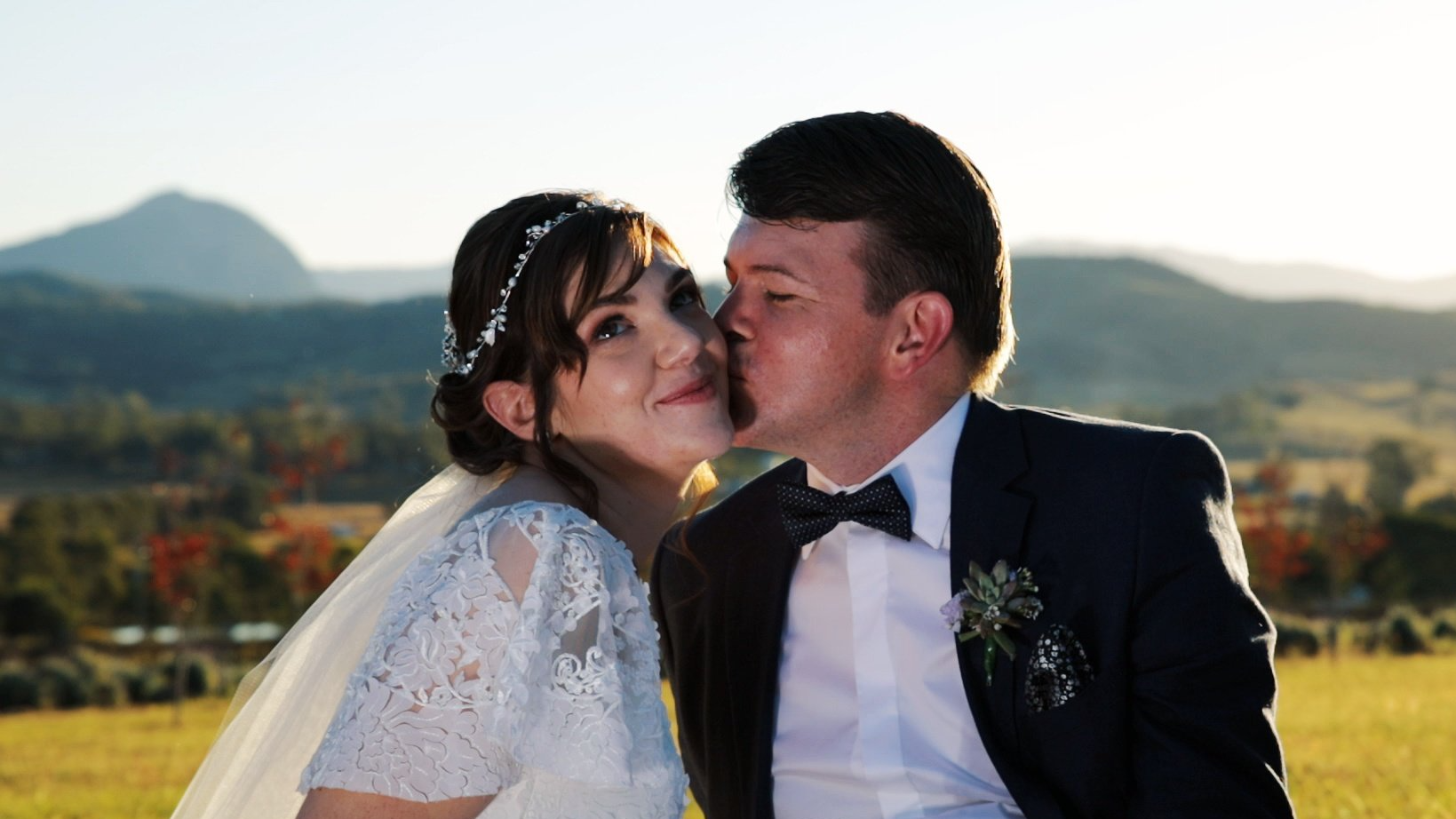 a bride and groom are kissing in a field with mountains in the background .
