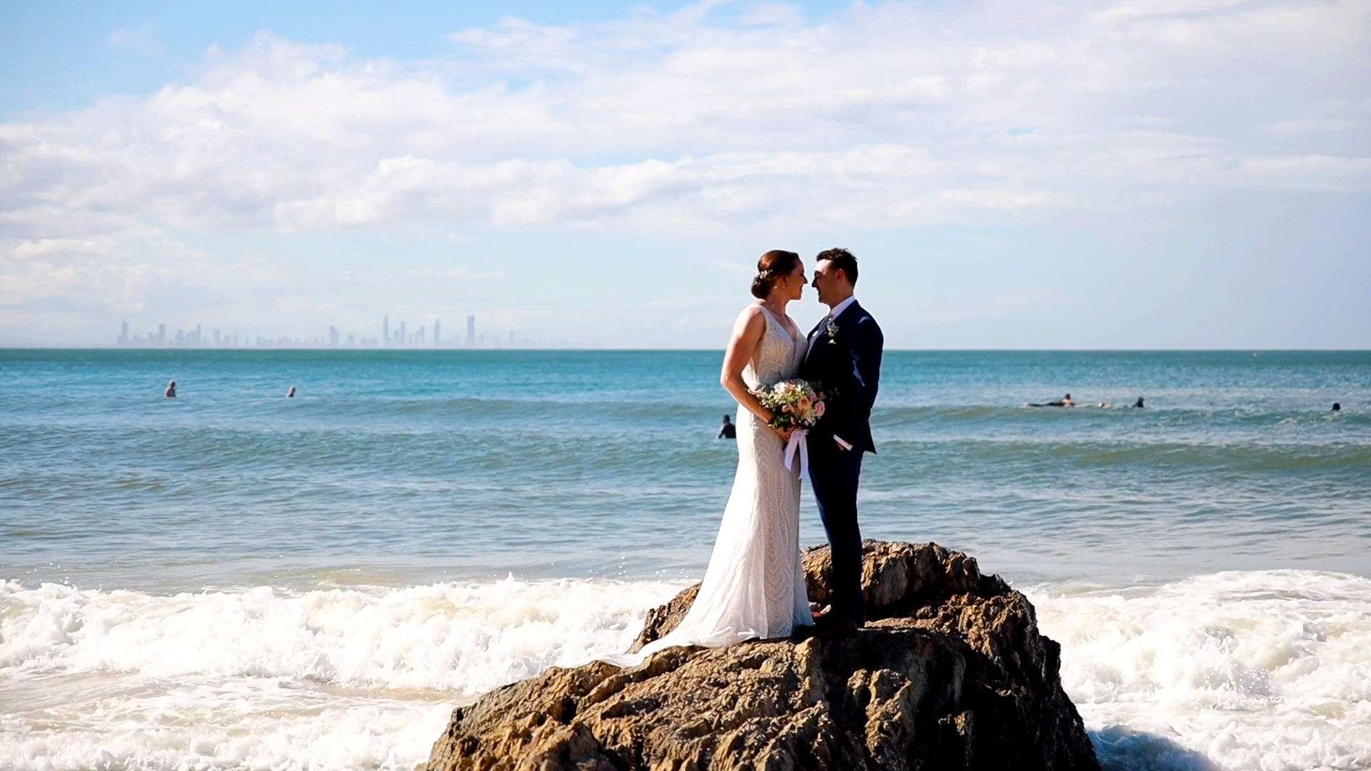 A bride and groom are standing on a rock near the ocean at the Gold Coast 