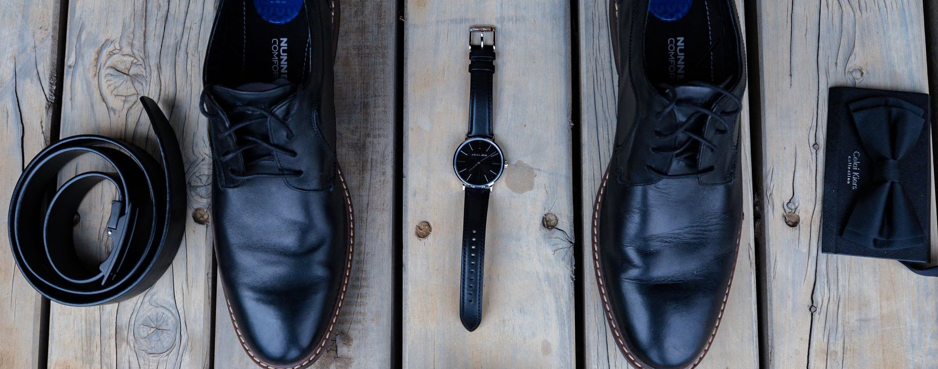 a pair of black shoes , a watch , a belt , and a bow tie are sitting on a wooden table .