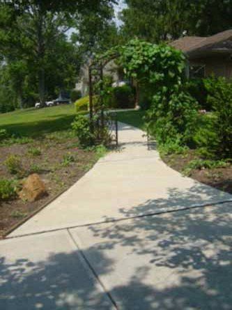 A concrete walkway leading to a house surrounded by trees and bushes.
