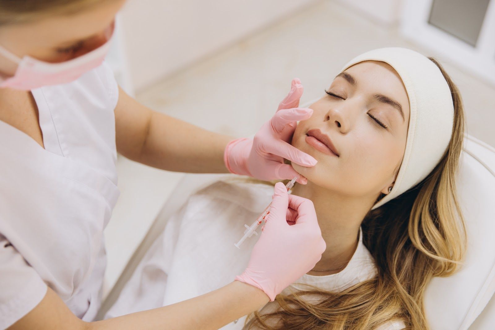 Cosmetician applying facial treatment to a relaxed client lying on a bed with a white headband