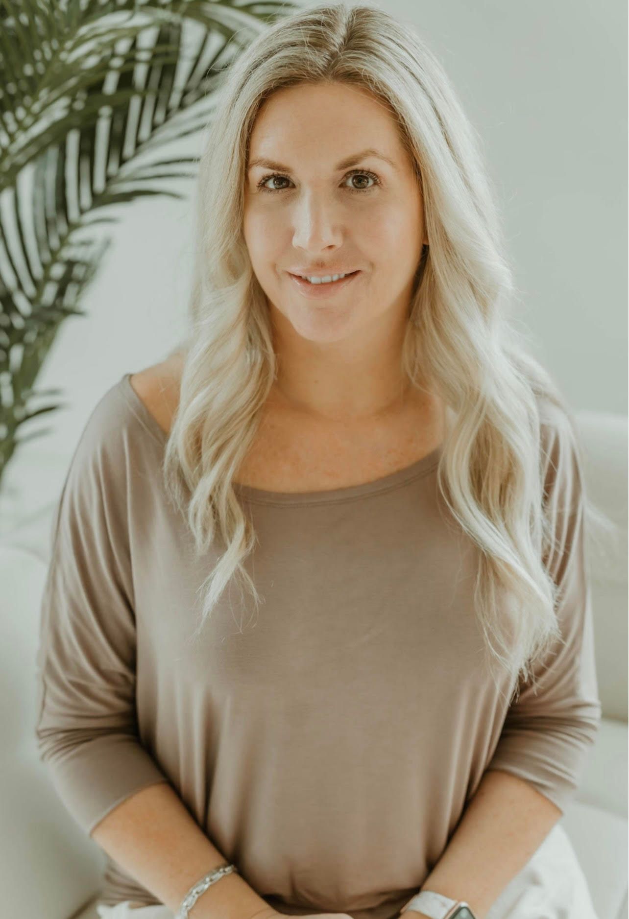 Blonde woman in a tan top standing indoors beside a leafy plant, facing the camera with a slight smile