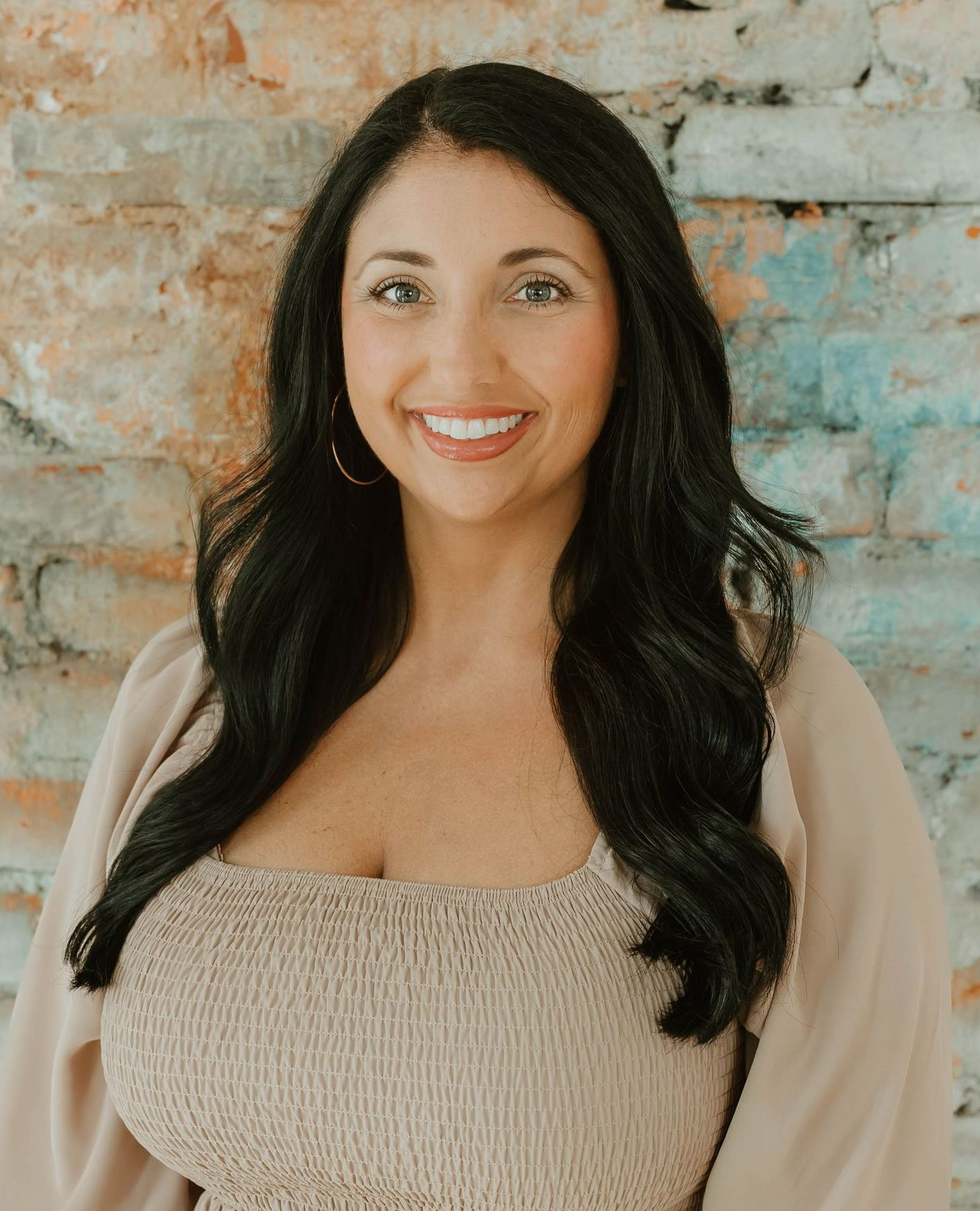 Smiling woman with long dark hair in a beige top, standing against a textured brick wall.