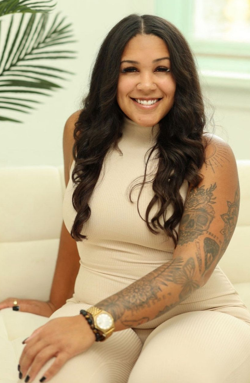 Smiling woman in a beige sleeveless outfit seated indoors beside a plant, with long dark hair and tattooed arm