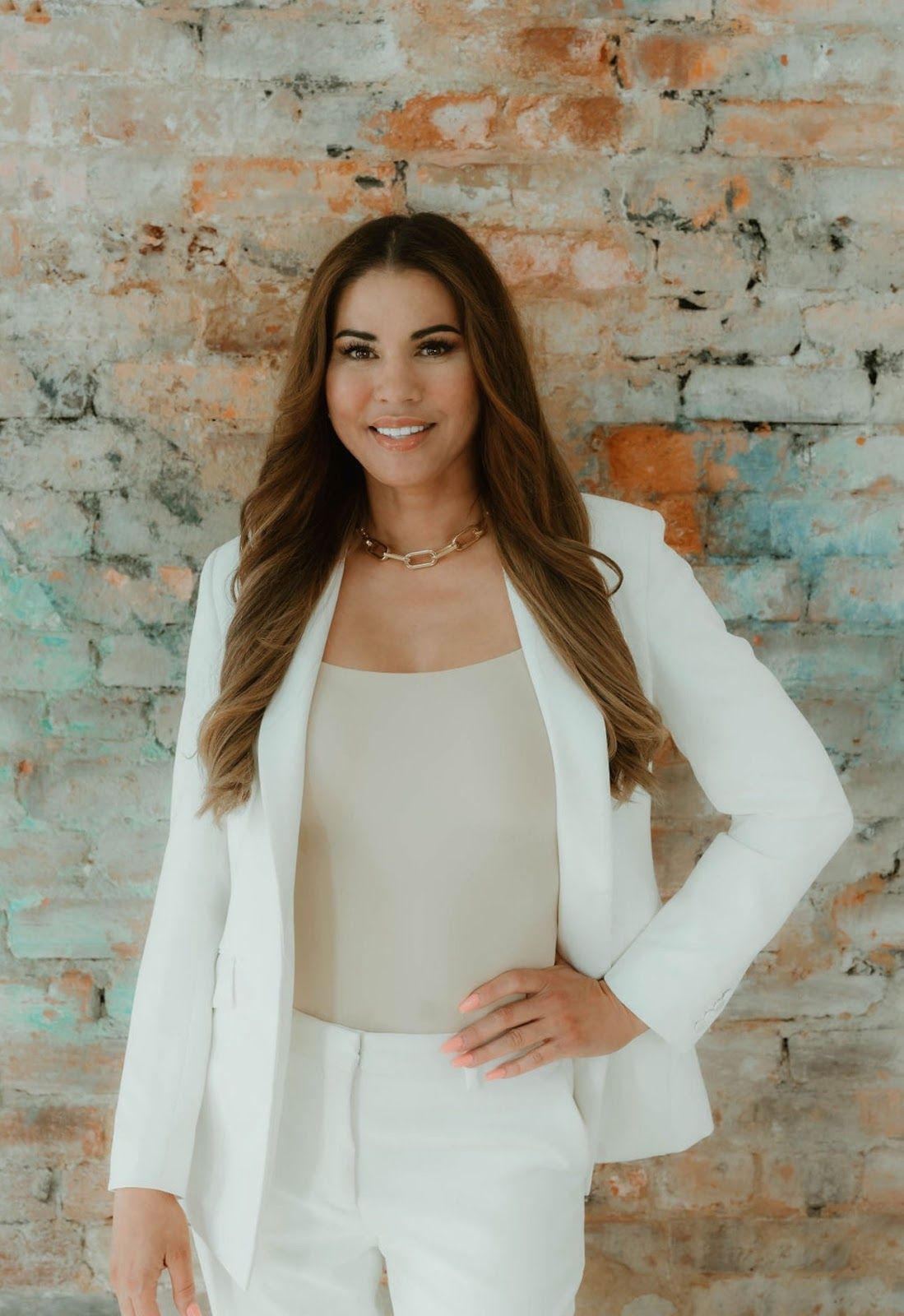 Woman in a white suit standing against a textured brick wall, smiling with one hand on her hip