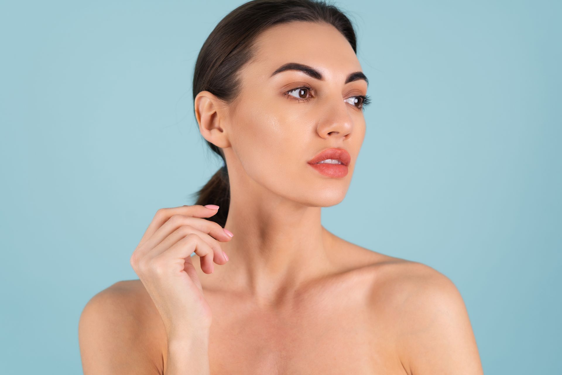 Woman with bare shoulders against a light blue background, looking to the side and touching her neck
