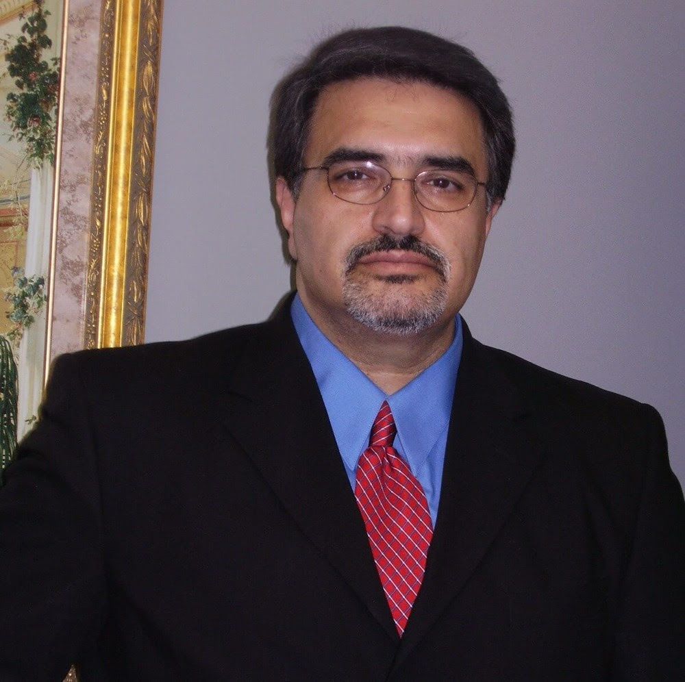 Man in a black suit, blue shirt, and red tie posing indoors beside a framed painting.