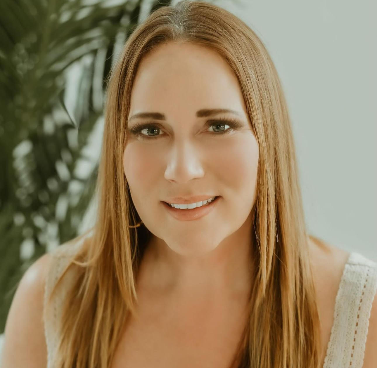 Smiling woman with long hair in a light sleeveless top, indoors with soft natural light and greenery behind her