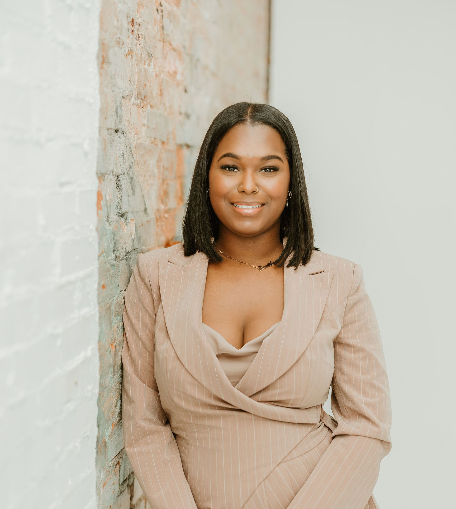 Woman in a tan blazer dress smiles, leaning against a brick wall in a white room.
