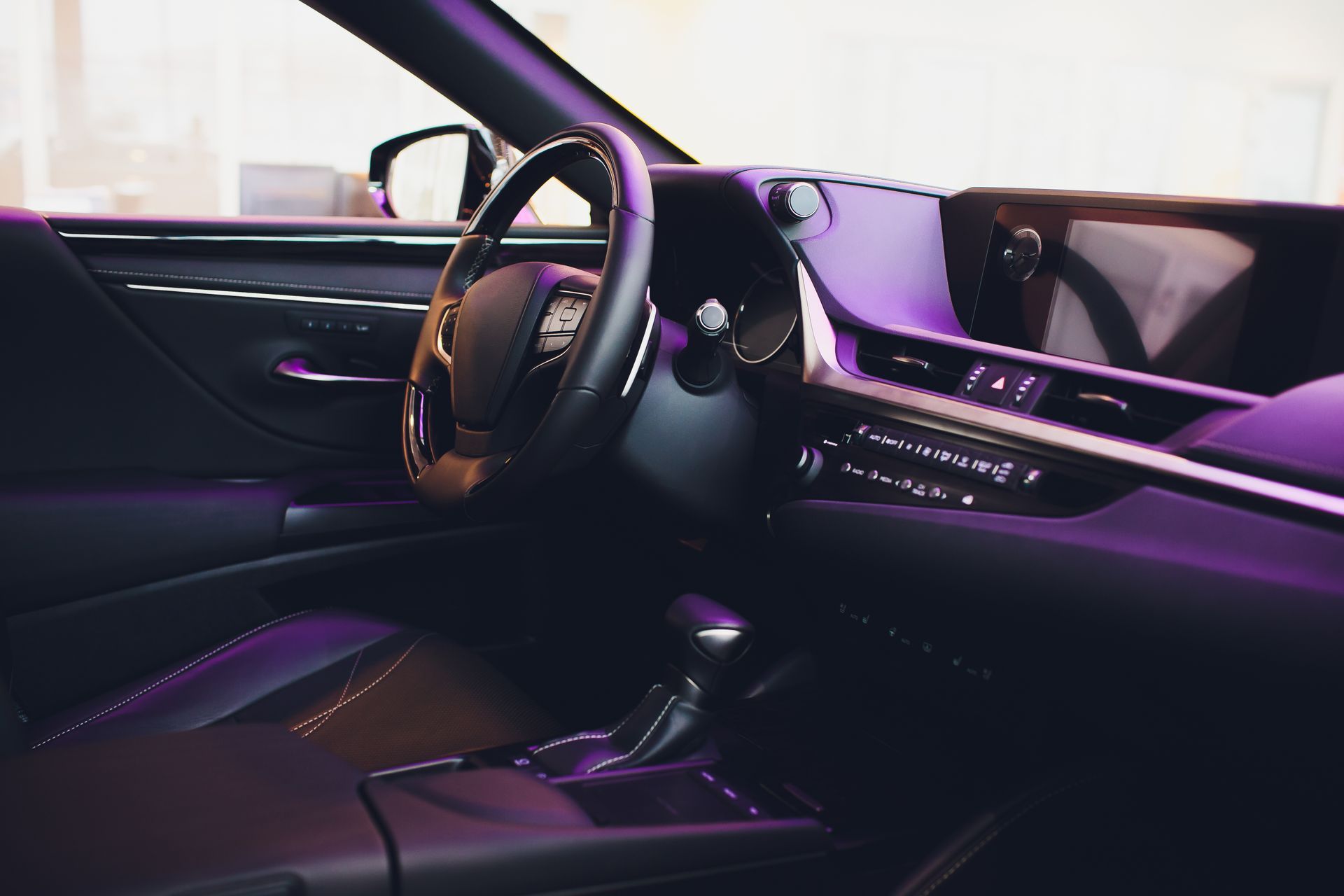 Black car interior with a leather steering wheel, dashboard, and gear shift. Purple lighting.