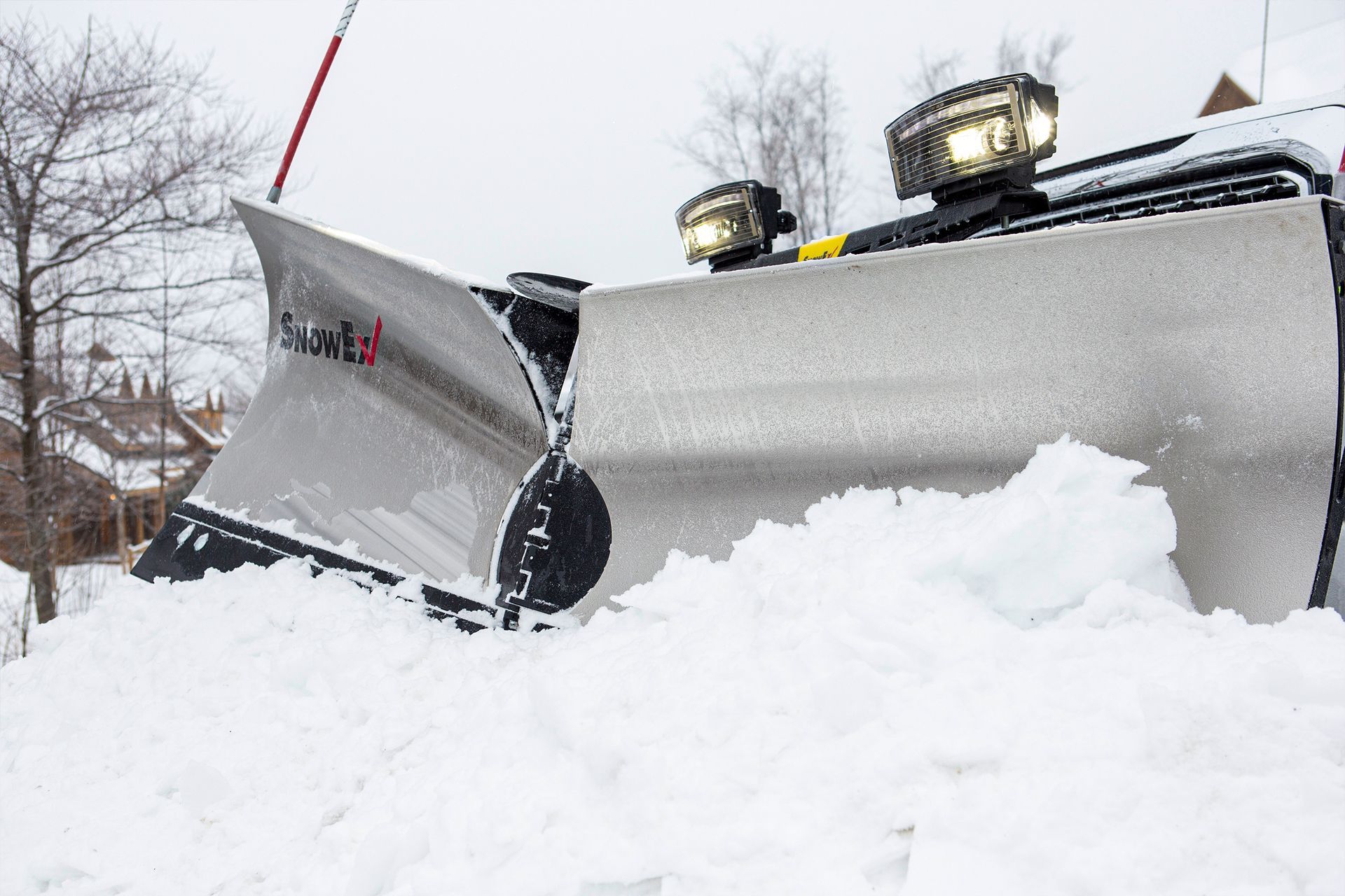 Snowplow clearing snow from a driveway, with bright lights illuminating the scene on a cloudy day.