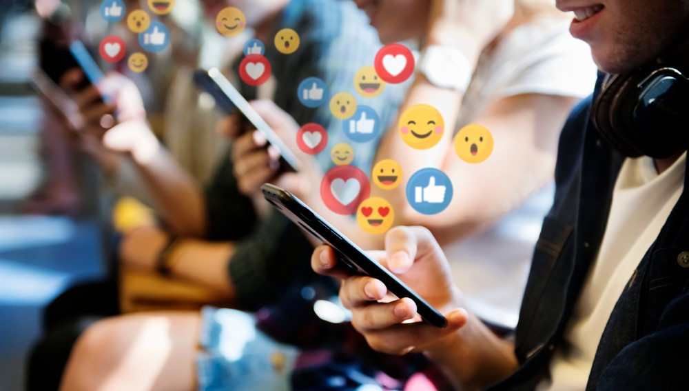 Plusieurs personnes tiennent des smartphones, entourés d'icônes de réactions de réseaux sociaux flottantes, comme des cœurs, des pouces levés et des émojis.